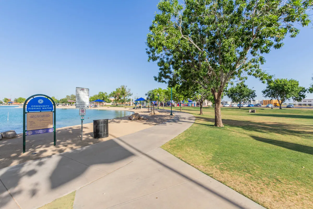 A lakeside park scene featuring a 'community fishing water' sign, a playground area, and a grassy area with trees. Children can be seen playing and walking paths invite leisurely strolls, making it a perfect spot for community and family gatherings.