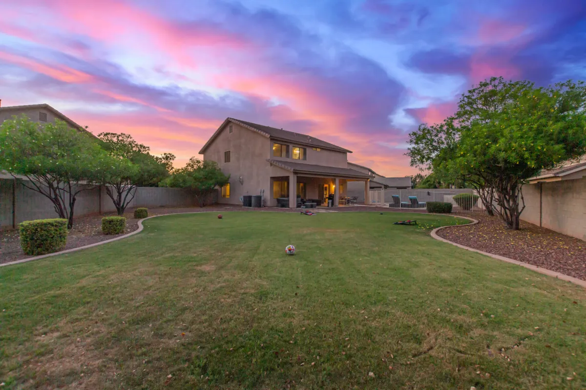 A charming view of the exterior of the house, surrounded by a lush, green lawn and mature trees. The sun is setting in the background, casting warm colors across the sky and illuminating the inviting patio area, perfect for gatherings or peaceful evenings outdoors.
