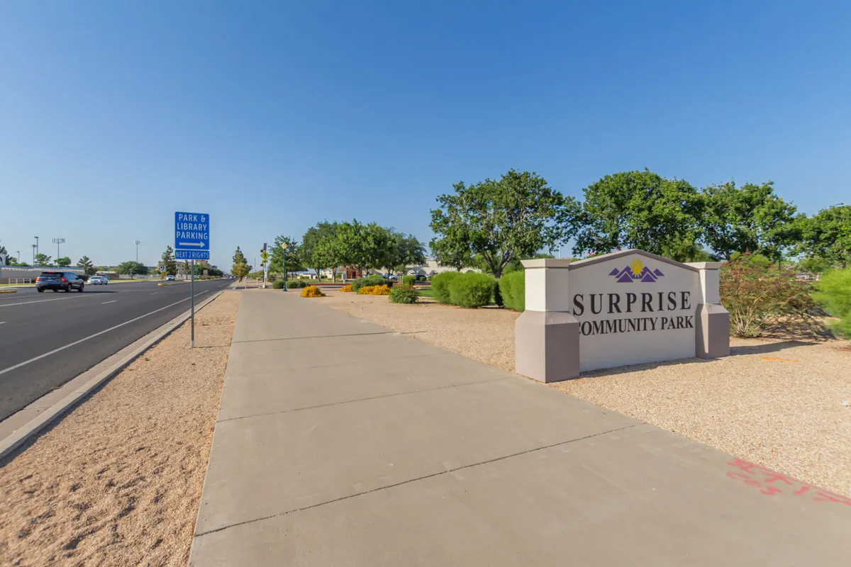 A welcoming view of the Surprise Community Park sign bordered by sidewalks and vibrant greenery, inviting visitors to explore recreational paths and enjoy family-friendly community events in a natural setting.
