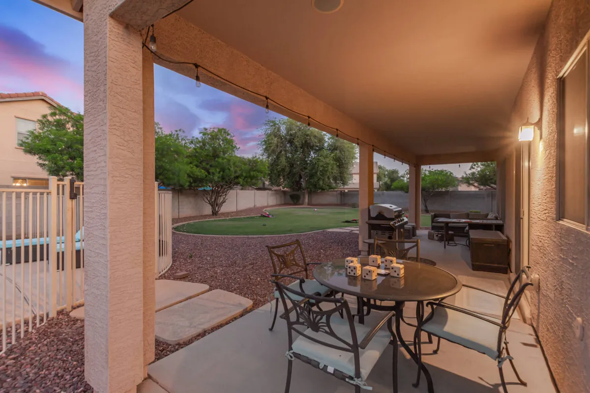 A table with candles is set on a patio next to a green lawn and a fenced pool area, with a grill nearby. The environment is casual and welcoming, ideal for outdoor dining, barbecuing, or enjoying games with family and friends as the sun sets.