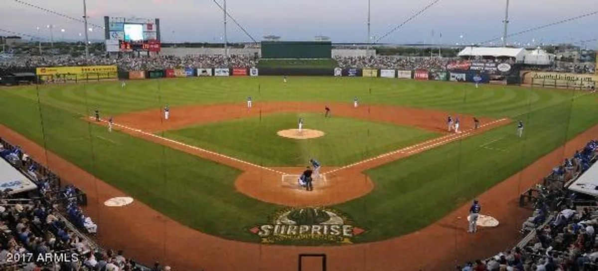 A vibrant baseball stadium scene showing players on the field, a crowd in the stands, and a visible scoreboard. The energy is festive, reflecting the excitement of a live sporting event.