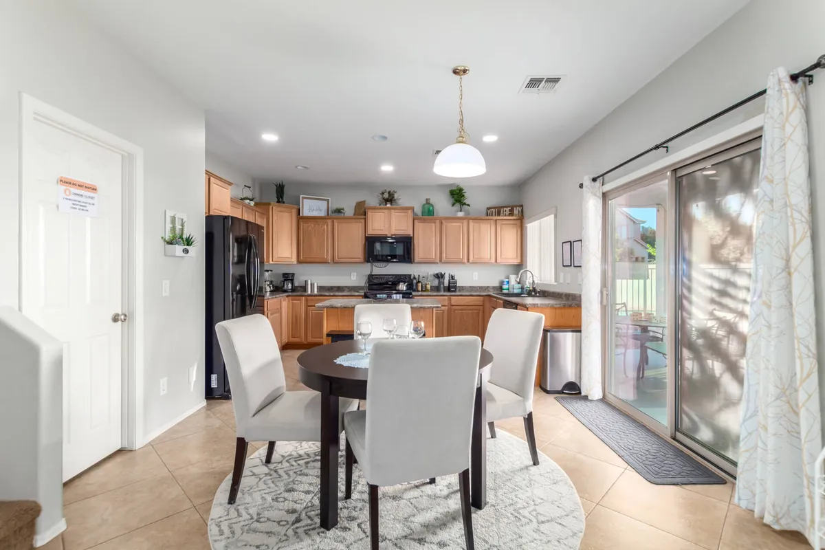 This image offers a view of the dining area with a spacious table, complemented by kitchen cabinets in the background and a sliding glass door that lets in natural light. The table is set up for family meals or casual gatherings, and the overall aesthetic is warm and inviting, with subtle desert-inspired touches.