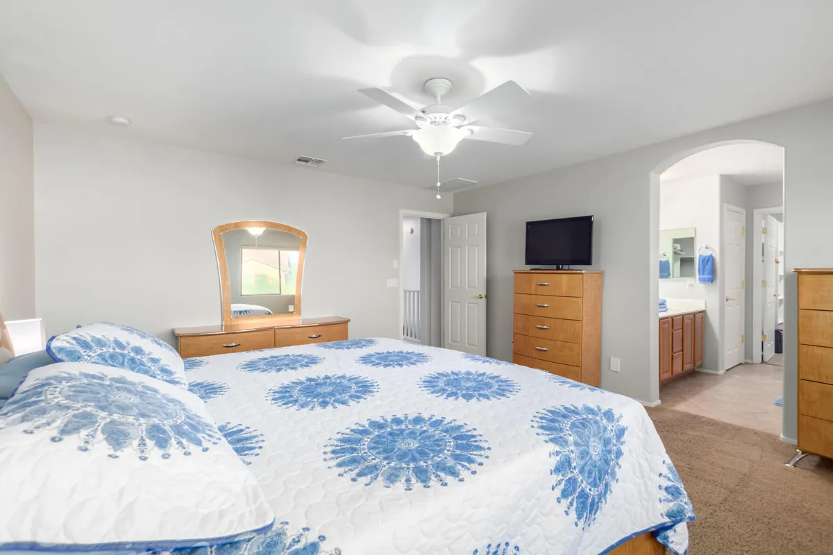 A serene bedroom featuring a bed with a blue and white patterned quilt, wooden dressers, a mirror, and a TV on the dresser. The space is softly lit, exuding comfort and tranquility, ideal for rest and relaxation.