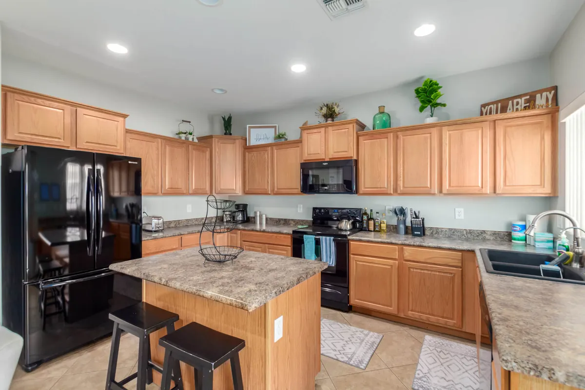 The photo features a welcoming kitchen with a prominent black refrigerator, a central kitchen island, wooden cabinetry, green plants adding a fresh touch, and decorative items that evoke a warm, lived-in atmosphere. The open layout suggests ample space for meal prep and family gatherings.