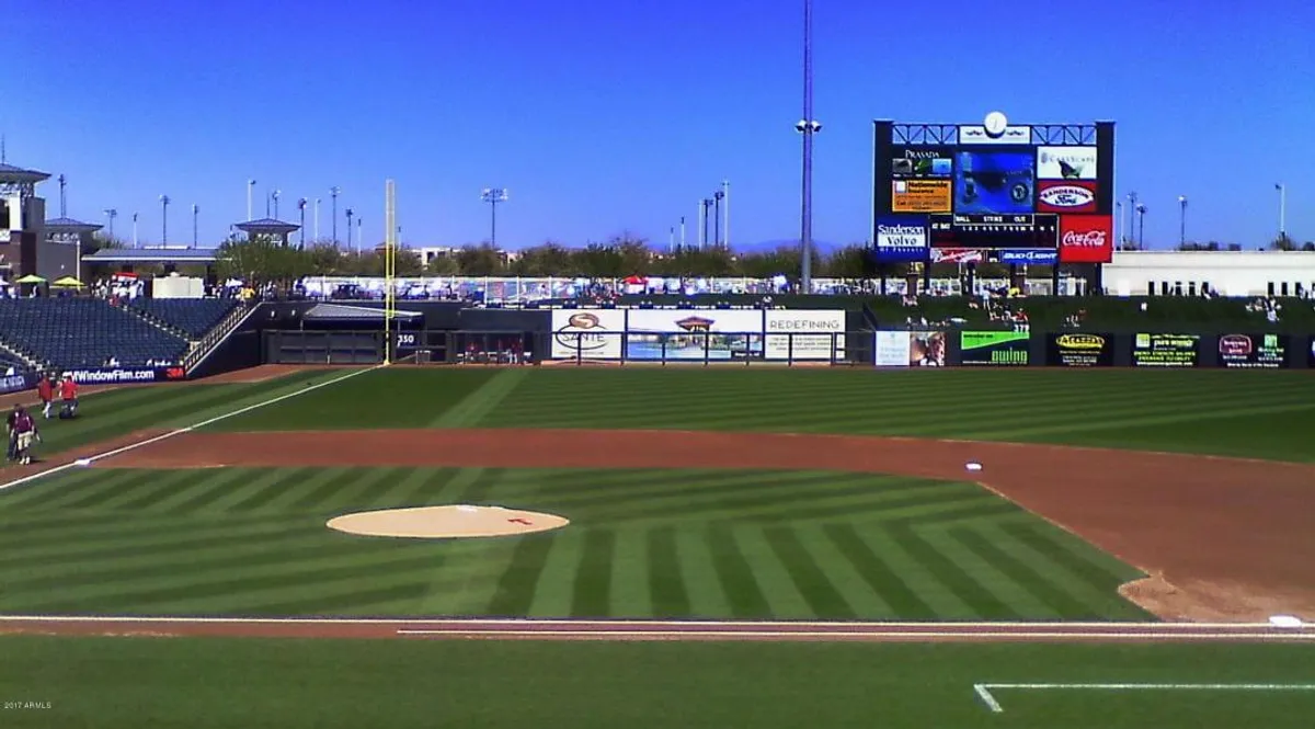 A bustling baseball stadium scene with a diamond, scoreboard, grandstands, and fans in the background. Spectators are engaged in the excitement, representing the thrill of catching a local game together as a family.